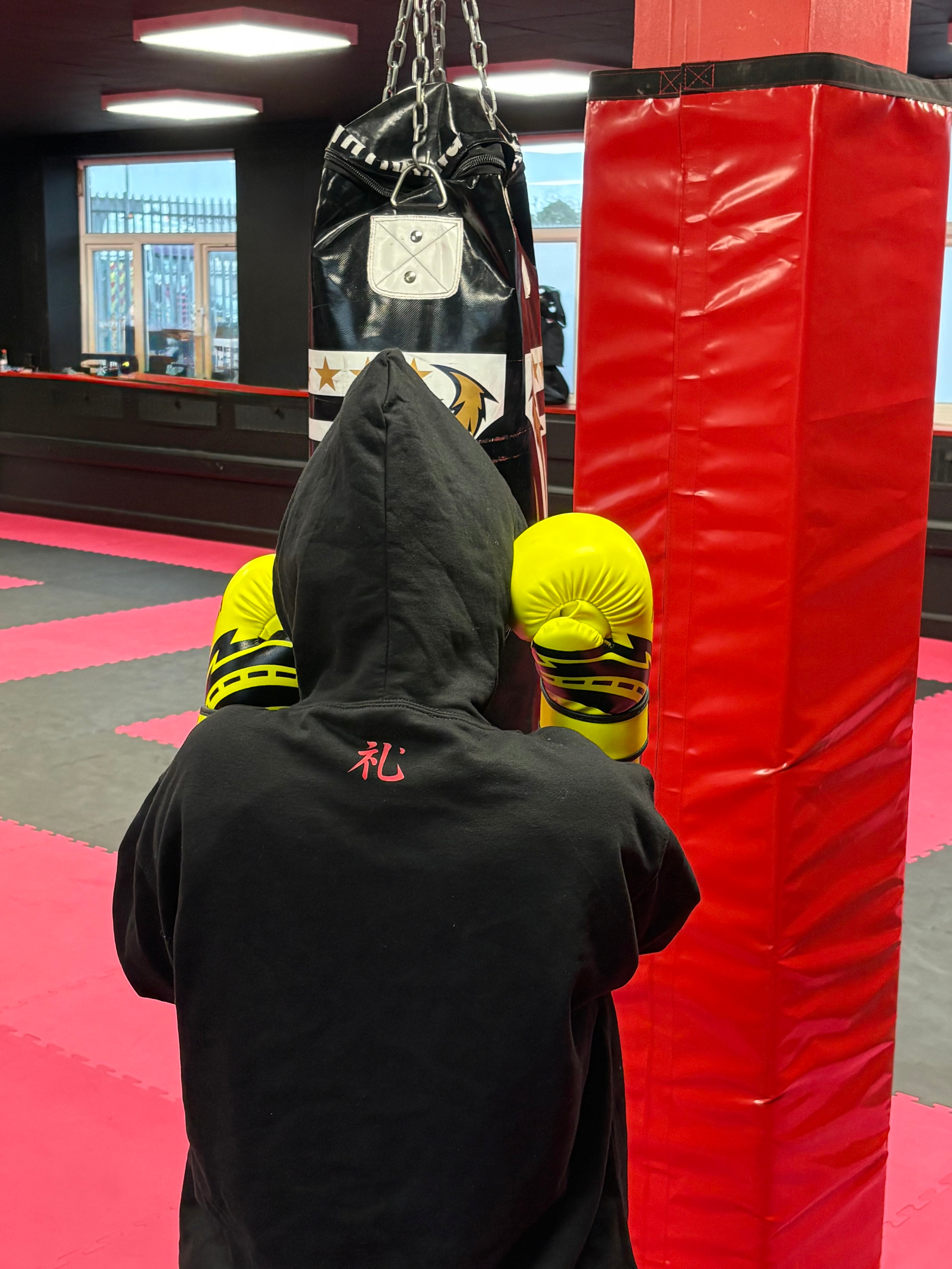 Person wearing a black hoodie and yellow headgear in a boxing ring with a punching bag.